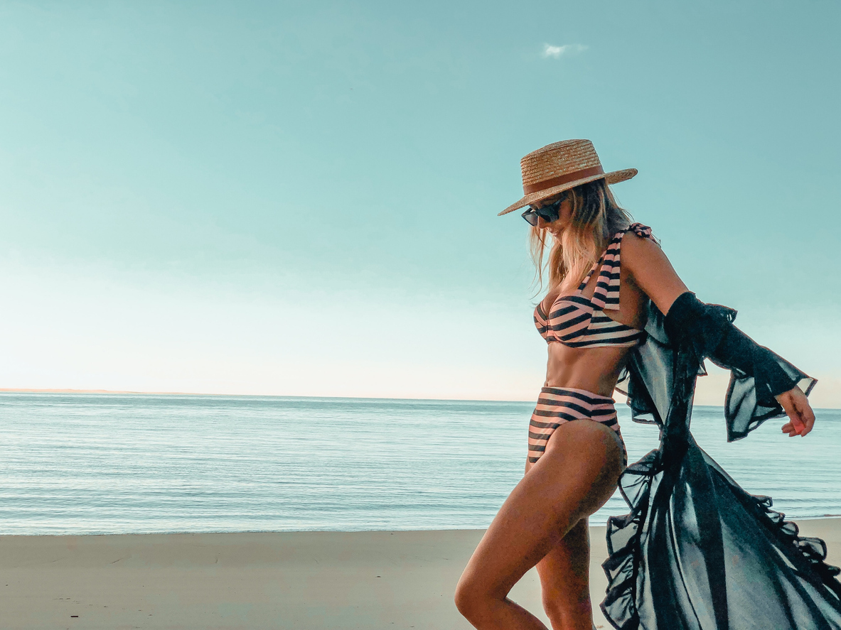 Beautiful woman at the beach in a stripped pinky bikini wearing a cover-up with mesh transparencies swimwear dress and a straw hat by the sea photography