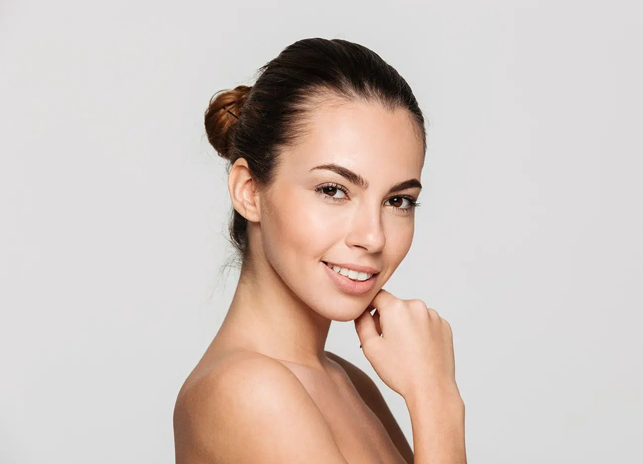 Young woman with hair in a bun gently touching her chin against a light grey background. Young woman with hair in a bun gently touching her chin against a light grey background.
